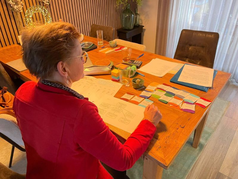 Ondernemer in rode jas werkt aan tafel aan haar plan, met kaartjes en papier voor zich